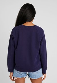 Tmavě modrá mikina s kulatým výstřihem a žebrovanými manžetami, s uvolněným střihem a měkkou texturou. Nošená s světle modrými džínovými šortkami.