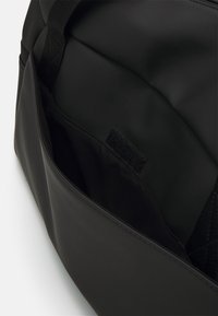 Sac de voyage noir avec une finition lisse et mate, un intérieur spacieux et une poche avec velcro à l'avant pour un accès facile.