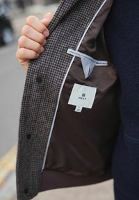 Hand holding open a brown checkered jacket revealing a smooth dark brown lining with a white NEXT brand label inside.