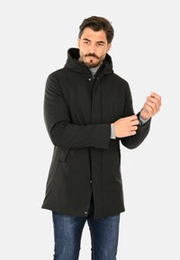 Giacca con cappuccio verde scuro, chiusura con bottoni a pressione, tasche laterali e design a mezza lunghezza. Realizzata in tessuto liscio e leggero.