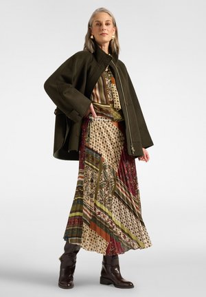 Mujer con cabello gris que lleva una falda estampada, top a juego, chaqueta verde oscuro, botas marrones y pendientes dorados, de pie con la mano en la cadera.