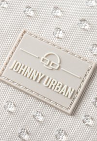 Un tissu blanc texturé présente des points en relief, avec une étiquette rectangulaire crème clair embossée "JOHNNY URBAN" en lettres grasses.