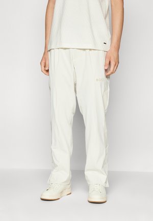 Person trägt weite weiße Hosen mit "BOSS"-Logo am Oberschenkel, weiße Turnschuhe und ein strukturiertes ärmelloses weißes Oberteil, steht vor einem weißen Hintergrund.