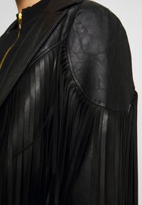 Czarna skórzana kurtka z długimi frędzlami na rękawach, ostrym kołnierzem i złotym zamkiem błyskawicznym z przodu.