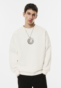 Mladý muž s krátkými tmavými vlasy, oblečený v bílém svetru, černých kalhotách a velkým kulatým stříbrným náhrdelníkem s fotografií uvnitř.