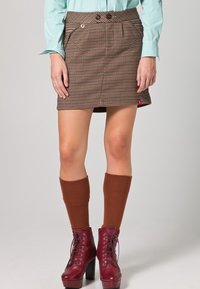 Brązowa mini spódnica w kratę z dwoma przednimi kieszeniami i guziczkami, zestawiona z brązowymi podkolanówkami i ciemnoczerwonymi, sznurowanymi butami na platformie.