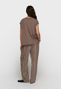 Mujer con cabello largo y oscuro, vistiendo una blusa marrón sin mangas y pantalones anchos con rayas verticales beige y negras, de pie con las manos en los bolsillos.