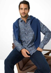 Blau gemustertes Hemd zum Knöpfen mit weißen und braunen Akzenten, kombiniert mit einem einfarbigen blauen Pullover, der über die Schultern gelegt ist. Dunkle Jeans.