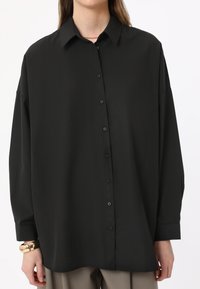 Black, long-sleeve, button-up shirt made of smooth fabric. Features a collar, front buttons, and slightly loose fit with a rounded hem.