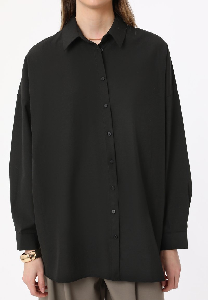 Black, long-sleeve, button-up shirt made of smooth fabric. Features a collar, front buttons, and slightly loose fit with a rounded hem.