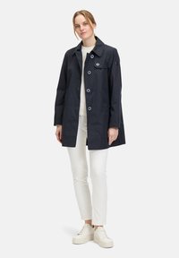 Marineblauer wasserdichter Mantel mit Knopfverschluss, großen Taschen und spitzem Kragen, kombiniert mit weißen Hosen und Sneakers.