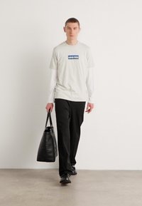 Lys grå T-skjorte med en blå logo på brystet, lagdelt over en hvit langermet skjorte, kombinert med svarte bukser og en svart totebag.