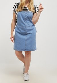 Džínová šaty s kšandami v světle modré barvě, s nastavitelnými popruhy, předními kapsami a přiléhavým živůtkem, v kombinaci s černobílým pruhovaným tričkem pod nimi.