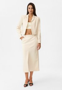 Blazer court beige clair avec fermeture à bouton, associé à une jupe midi assortie avec une fente sur le côté. Porté avec des talons noirs à lanières.