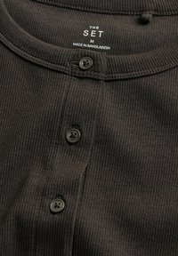 Dunkelbraunes, geripptes Oberteil mit drei schwarzen Knöpfen und Etikett "THE SET M Made in Bangladesh" im Kragen.