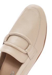 Mocassino in pelle beige con dettaglio di cucitura sulla parte superiore e una texture liscia, dotato di una suola marrone chiaro. L'interno è morbido.