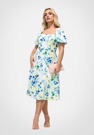 Mujer con un vestido midi blanco con estampado floral azul, mangas abullonadas, escote cuadrado, sosteniendo un clutch rosa y llevando tacones nude con tiras en el tobillo.