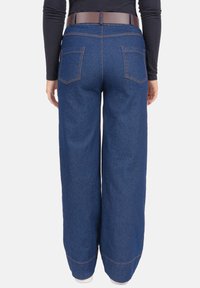Pantalon large en denim, bleu foncé, avec surpiqûres contrastantes et poches arrière. Associé à une ceinture marron, à la texture lisse.