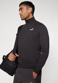 Sweat à zip noir avec col montant, arborant un petit logo Puma blanc sur la poitrine, associé à un pantalon de sport sombre et un sac noir.