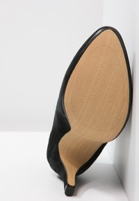 Zapato de tacón alto de ante negro con punta afilada, que presenta una suela beige suave con un patrón texturizado para mayor agarre.