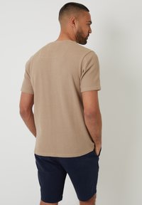 Beige strukturiertes T-Shirt mit kurzen Ärmeln und Rundhalsausschnitt, kombiniert mit marineblauen Shorts. Das Material wirkt weich und leicht. Sichtbare Nähte.