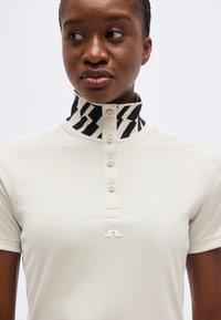 Γυναίκα που φοράει λευκό πόλο μπλουζάκι με κουμπιά και ψηλό ασπρόμαυρο σχέδιο στο γιακά, κοιτάζοντας ελαφρώς προς το πλάι.