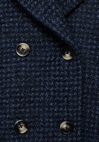 Mornarsko moder dvoredni plašč iz tvida, ki se ponaša z teksturirano blago, ovratnikom z zarezo in dvema okroglima črnima gumboma z vzorcem križa.