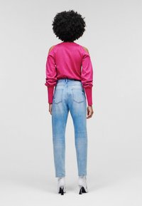 Frau mit lockigem Haar, die einen leuchtend pinken Cold-Shoulder-Pullover, hellblaue Jeans und weiße, knöchelhohe Stiefel mit Absatz trägt, und mit dem Rücken zu sehen ist.