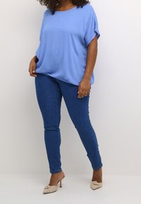 Blusa azul claro de corte holgado con mangas cortas, combinada con vaqueros ajustados azul oscuro y mules beige adornadas con nudos.