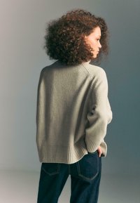 Donna con capelli ricci che indossa un maglione grigio chiaro e jeans blu scuri, in piedi con la schiena rivolta e le mani in tasca.