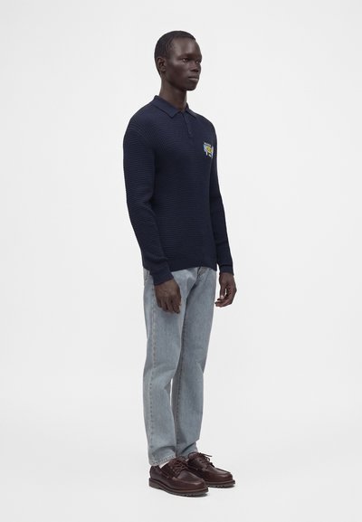 Homme debout portant un polo en maille bleu foncé à manches longues, un jean bleu clair et des chaussures en cuir marron foncé sur un fond blanc uni.