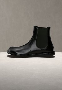 Botte Chelsea en cuir noir avec panneaux latéraux élastiques et petit talon empilé sur une surface en pierre claire avec un fond gris.