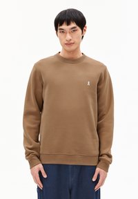 Man in een bruine lange mouwen sweater met een klein wit logo op de linkerborst en donkere blauwe broek tegen een effen witte achtergrond.