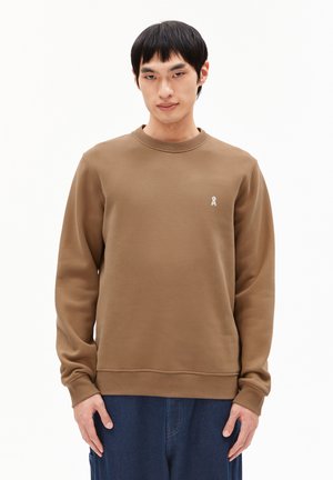 Man in een bruine lange mouwen sweater met een klein wit logo op de linkerborst en donkere blauwe broek tegen een effen witte achtergrond.