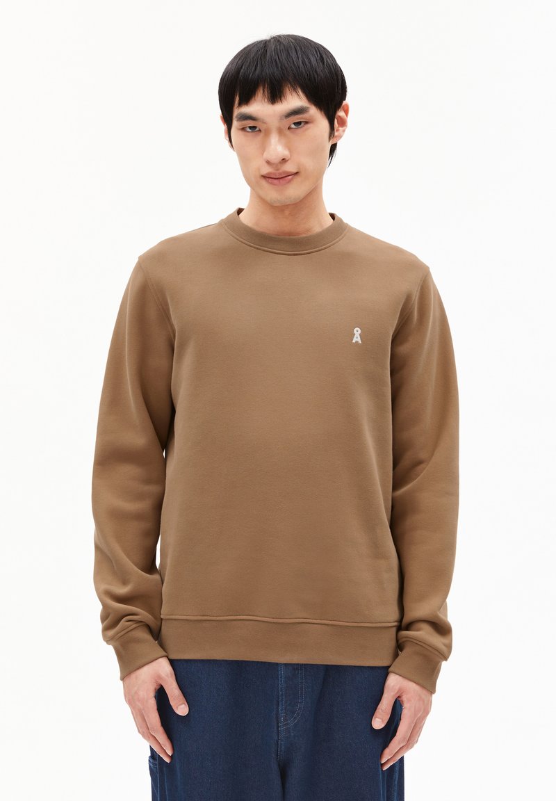 Man in een bruine lange mouwen sweater met een klein wit logo op de linkerborst en donkere blauwe broek tegen een effen witte achtergrond.