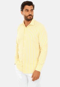 Camicia a righe gialle e bianche con colletto, maniche lunghe e bottoni bianchi. Il tessuto sembra liscio e leggero.