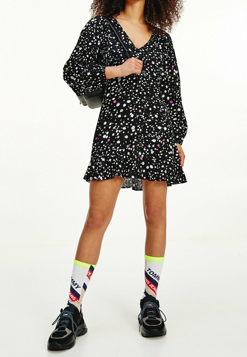Femme portant une robe noire à fleurs à col en V, des chaussettes blanches avec logo, des baskets noires, et un sac à épaule noir.