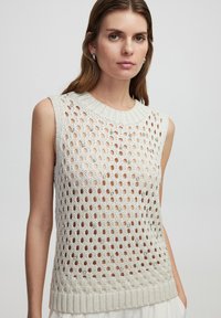 Top de punto sin mangas con patrones circulares abiertos en beige claro. Cuello y dobladillo acanalados. Material suave y texturizado.