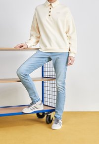 Sudadera color crema con tapeta de botones y logo bordado, combinada con jeans ajustados de color azul claro y zapatillas grises con rayas.