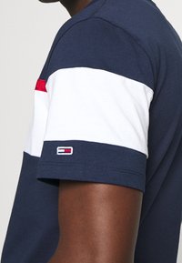 Osoba s tmavou pletí oblečená v krátkém rukávu námořnické modré a bílé košili s malým červeným, bílým a modrým logem na rukávu.