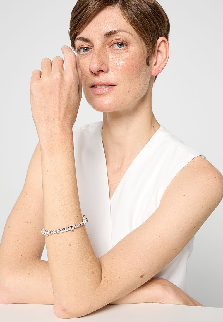 Silberne Kettenarmband mit miteinander verbundenen Gliedern, mit einem Verschluss, getragen an einem nackten Handgelenk vor einem schlichten Hintergrund.