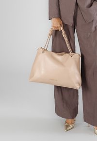 Beige tote taske lavet af glat læder med et flettet håndtag og hardware i guld tone. Rummelig design og subtil branding på forsiden.