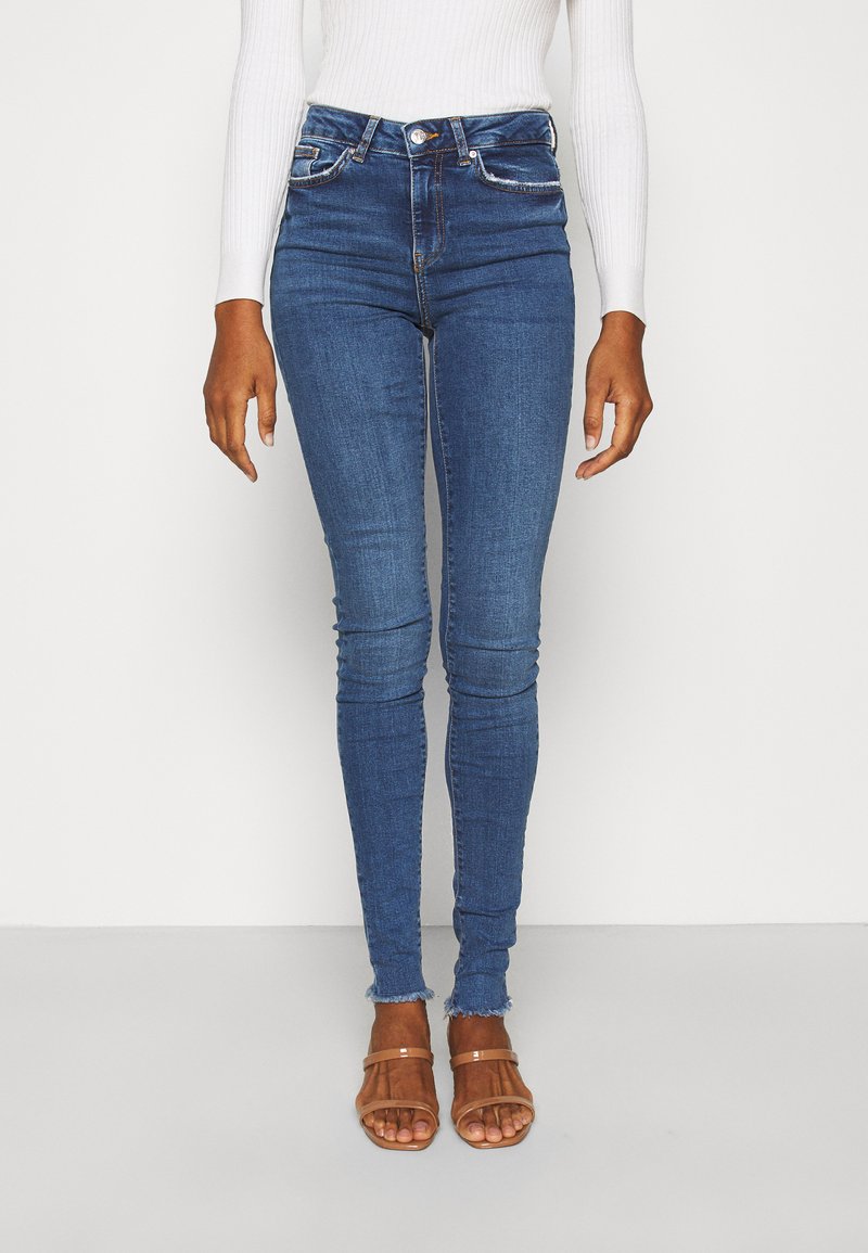 Calças skinny azuis feitas de denim com uma cintura alta, apresentando bolsos frontais, desgaste subtil e barra desfiada, combinadas com sandálias cor de camelo.