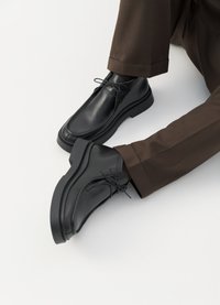 Svarta läderskor med rund tå, framträdande snörning och en tjock sula. Bärs med bruna, smala byxor som visar upp en elegant design.