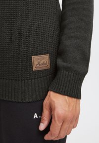 Dark green knit sweater with ribbed cuffs and hem. Features a brown label with "Solid" branding. Worn with black sweatpants showing white lettering.