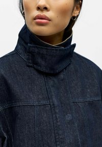 Giacca di denim scuro con collo alto, caratterizzata da un design strutturato e cuciture evidenti. Texture liscia, accenti minimi.