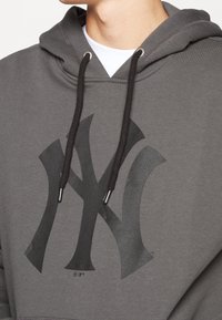 Sweat à capuche gris avec un grand logo NY noir, cordons ajustables avec embouts argentés, et une texture douce et lisse.