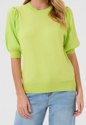 Frau trägt einen kurzärmligen Pullover in Limettengrün mit Puffärmeln und hellblaue Jeans, steht vor einem schlichten Hintergrund.