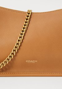 Geantă de piele maro deschis cu o textură netedă, dotată cu o curea din lanț auriu și logo-ul gravat "COACH NEW YORK" în partea de jos.