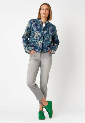 Woman standing with hands in pockets wearing blue floral button-up jacket, light gray cropped pants, and bright green sneakers.